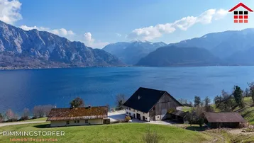 Anwesen mit mehreren Gebäuden direkt am Seeufer, umgeben von einer beeindruckenden Berglandschaft unter blauem Himmel.