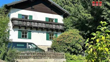 Traditionelles Haus mit Holzbalkon und grünen Fensterläden, umgeben von üppiger Natur.