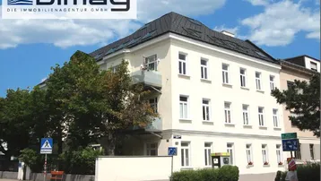 Mehrfamilienhaus mit heller Fassade, Mansarddach und kleinem Balkon an einer Straßenecke.