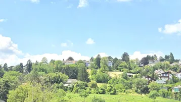 Weitläufiger Blick über eine grüne Hügellandschaft mit vereinzelten Häusern und viel Baumbestand.