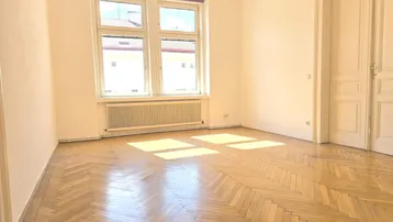 Helles Zimmer mit Fischgrätparkett und großen Fenstern, das viel Tageslicht hereinlässt.