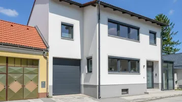 Modernes weißes Haus mit dunklen Fenstern und integrierter Garage an einer ruhigen Straße.