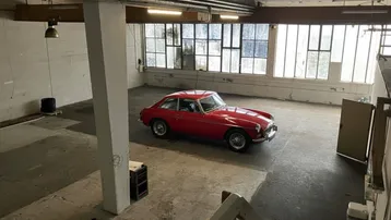 Großer, leerer Industrieraum mit Betonboden, hohen Fenstern und sichtbaren Leitungen, inklusive eines roten Oldtimers.