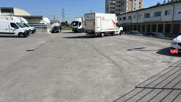 Großer asphaltierter Parkplatz mit Lieferwagen und Lastwagen, umgeben von Industriegebäuden und einem Wohnhochhaus unter blauem Himmel.