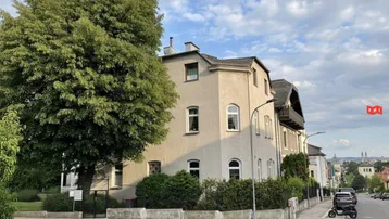 Ansicht eines Mehrfamilienhauses an einer Straßenecke mit altem Baumbestand und gepflegter Hecke.