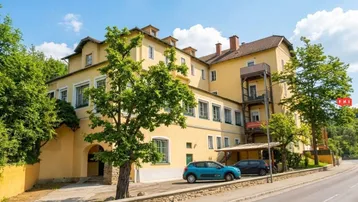 Gelbes Mehrfamilienhaus mit mehreren Etagen, vielen Fenstern und einem Carport an der Seite.