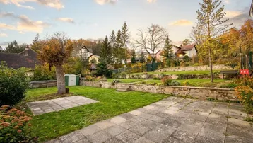 Großer, terrassierter Garten mit Rasenflächen, gepflasterter Terrasse und Gartenhaus, umgeben von Bäumen und Häusern.