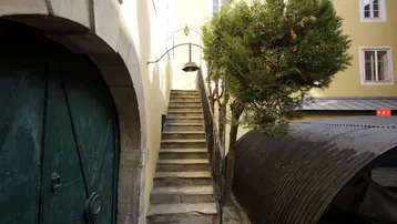 Eine alte Steintreppe führt zu einem oberen Eingang, umgeben von einer gelben Fassade und einem Baum.