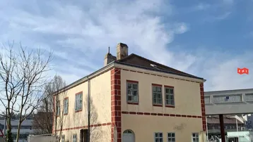 Historisches Gebäude mit beiger Fassade, roten Zierelementen und einem kleinen Außenbereich mit Sitzgelegenheiten unter blauem Himmel.