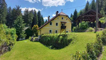 Gelbes Landhaus mit rotem Ziegeldach, umgeben von grünen Wiesen und hohen Bäumen unter blauem Himmel.