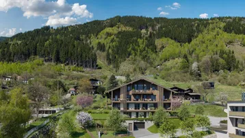 Modernes Gebäude im Chalet-Stil mit Balkonen und Tiefgaragenzufahrt, umgeben von grünen Hügeln und Bergen.