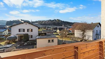 Blick vom Balkon auf die umliegende Wohnsiedlung und bewaldete Hügel unter blauem Himmel.