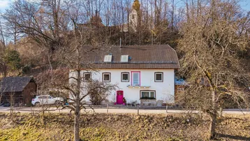 Weißes Haus mit rotem Dach und auffälliger pinker Tür, umgeben von Bäumen und Hügeln.