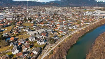 Luftaufnahme der Umgebung mit Wohngebäuden, Fluss und Bergen im Hintergrund.