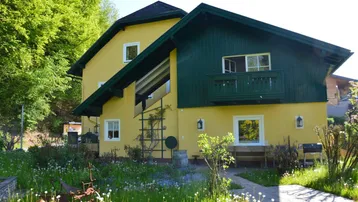 Gelbes Haus mit grünem Holzbalkon und Gartenbereich in ländlicher Umgebung.