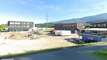 Baustelle mit modernen Gebäuden im Bau, umgeben von grüner Landschaft und Bergen.