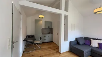 Heller Wohnbereich mit Holzboden, gemütlichem Sofa und integrierter Küchenzeile unter Dachschrägen.