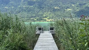 Ein Holzsteg führt durch hohes Schilf direkt zum See mit beeindruckendem Bergblick.