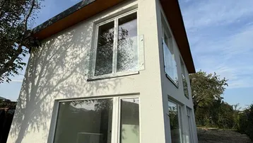 Moderne Hausfassade mit großen Fenstern, weißem Putz und Glasgeländer unter blauem Himmel.