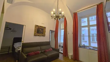 Gemütlicher Wohnbereich mit Sofa, kleiner Küchenzeile, Kronleuchter und eleganten roten Vorhängen.