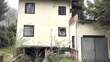 Altes Haus mit beiger Fassade, kleinem Balkon und integrierter Garage, umgeben von Bäumen und Sträuchern.