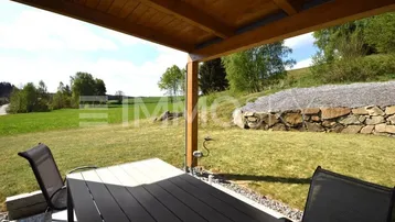 Überdachte Terrasse mit Esstisch und Stühlen, umgeben von grüner Landschaft und Steinmauer.