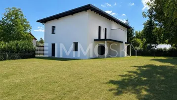 Modernes Einfamilienhaus mit weißer Fassade, großem Rasen und umgebenden Bäumen unter blauem Himmel.
