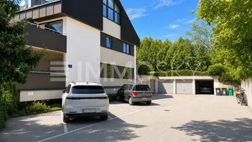 Mehrfamilienhaus mit Parkplätzen und Garagen in grüner Umgebung unter blauem Himmel.
