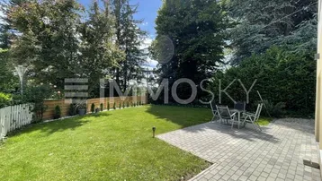 Gepflegter Gartenbereich mit großer gepflasterter Terrasse und Sitzgelegenheiten im Freien, umgeben von Bäumen.