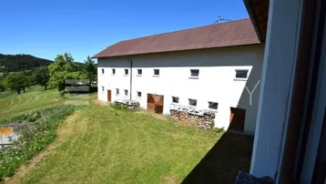 Langes, weißes Gebäude mit braunem Dach und Holzstapeln, umgeben von grünen Hügeln.