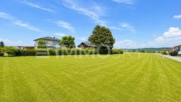 Weitläufige grüne Wiese mit Einfamilienhäusern im Hintergrund unter blauem Himmel.