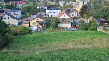 Blick über eine ländliche Wohnsiedlung mit Einfamilienhäusern und grünen Hügeln unter blauem Himmel.