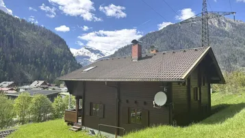 Traditionelles Holzhaus mit Satteldach und Bergblick, umgeben von grünen Wiesen und Bäumen.