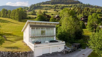 Modernes weißes Haus mit großen Fensterfronten, Balkonen und einer möblierten Terrasse in grüner Hanglage.