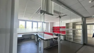 Modernes Büro mit Glaswänden, großen Fenstern und Blick ins Grüne, ausgestattet mit Schreibtischen und Regalen.