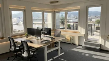 Modernes Büro mit großen Fenstern, viel Tageslicht und direktem Zugang zu einer Terrasse mit Stadt- und Bergblick.
