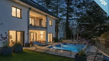 Abendliche Ansicht einer modernen Villa mit beleuchtetem Pool, Garten und Balkon.