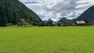 Weite grüne Wiese mit malerischem Bergblick und vereinzelten Häusern im Hintergrund unter bewölktem Himmel.