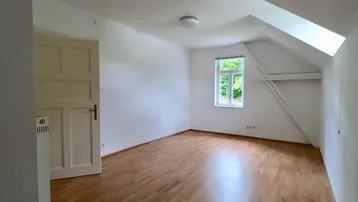Heller Raum im Dachgeschoss mit Holzboden, Dachschräge und einem Fenster mit Grünblick.