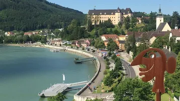 Panoramablick auf einen See, ein malerisches Dorf und ein historisches Schloss unter blauem Himmel.