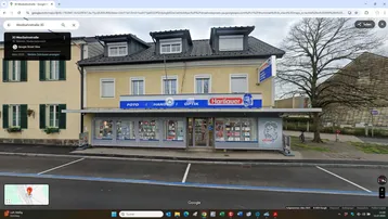 Gelbes Gebäude mit Geschäftsfassade und Dachgauben, aufgenommen von der Straße aus mit Parkplätzen davor.