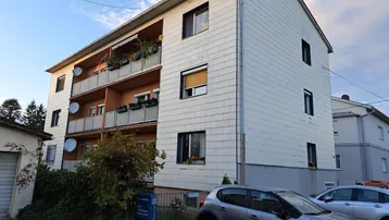 Mehrfamilienhaus mit mehreren Balkonen, weißer Fassadenverkleidung und Satteldach unter blauem Himmel.