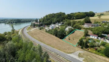 Luftaufnahme eines Grundstücks am Flussufer mit angrenzender Straße und Bahnlinie unter klarem Himmel.
