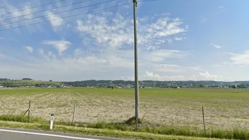Weitläufige Landschaft mit Feldern, einem Strommast und einem Dorf in der Ferne unter blauem Himmel.