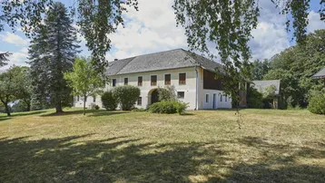 Großes Landhaus mit weitläufigem Rasen und altem Baumbestand unter blauem Himmel.