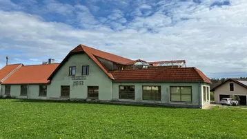 Gebäude mit hellgrüner Fassade und rotem Ziegeldach, umgeben von einer grünen Wiese unter bewölktem Himmel.