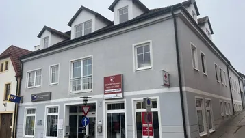 Graues Mehrfamilienhaus mit weißer Fassade und Dachgauben an einer Straße gelegen, mit Geschäftsräumen im Erdgeschoss.