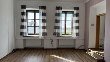 Leerer Raum mit zwei Fenstern, Laminatboden und Heizkörpern unter den Fenstern, gut beleuchtet.