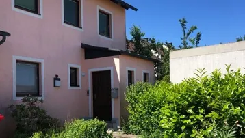 Hausfassade in Rosa mit Vorgarten und Hecke unter blauem Himmel.