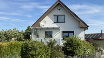Vorderansicht des Einfamilienhauses mit verglastem Balkon und gepflegtem Vorgarten unter blauem Himmel.